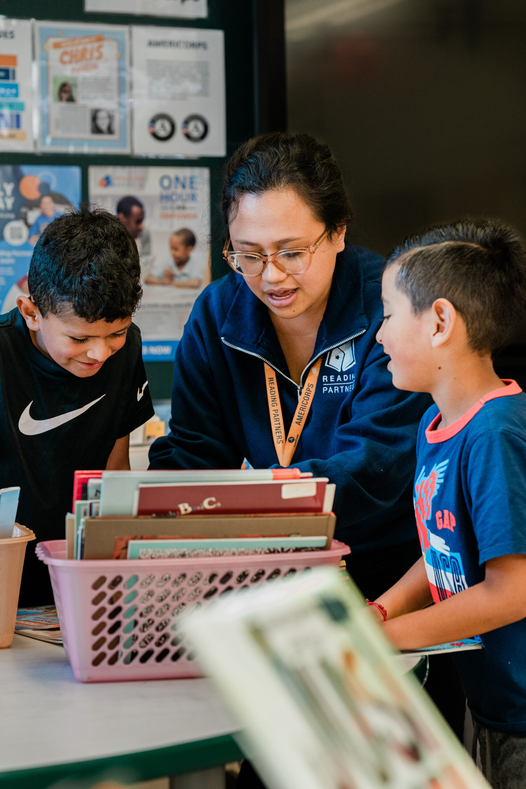 A teacher assists two young boys with their reading at a table filled with books in a classroom.