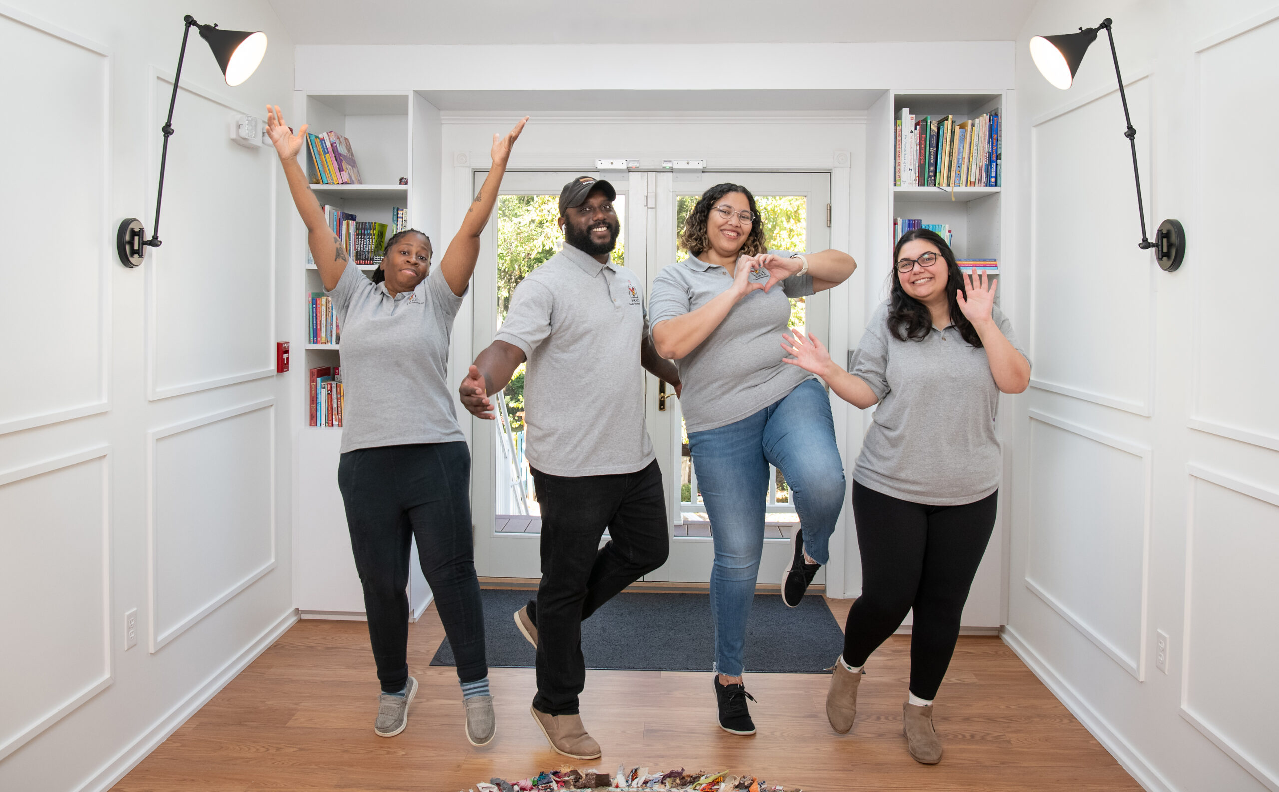 Four people in grey shirts pose enthusiastically in a well-lit room with bookshelves. Two have arms raised, one forms a heart with their fingers, and the other waves.