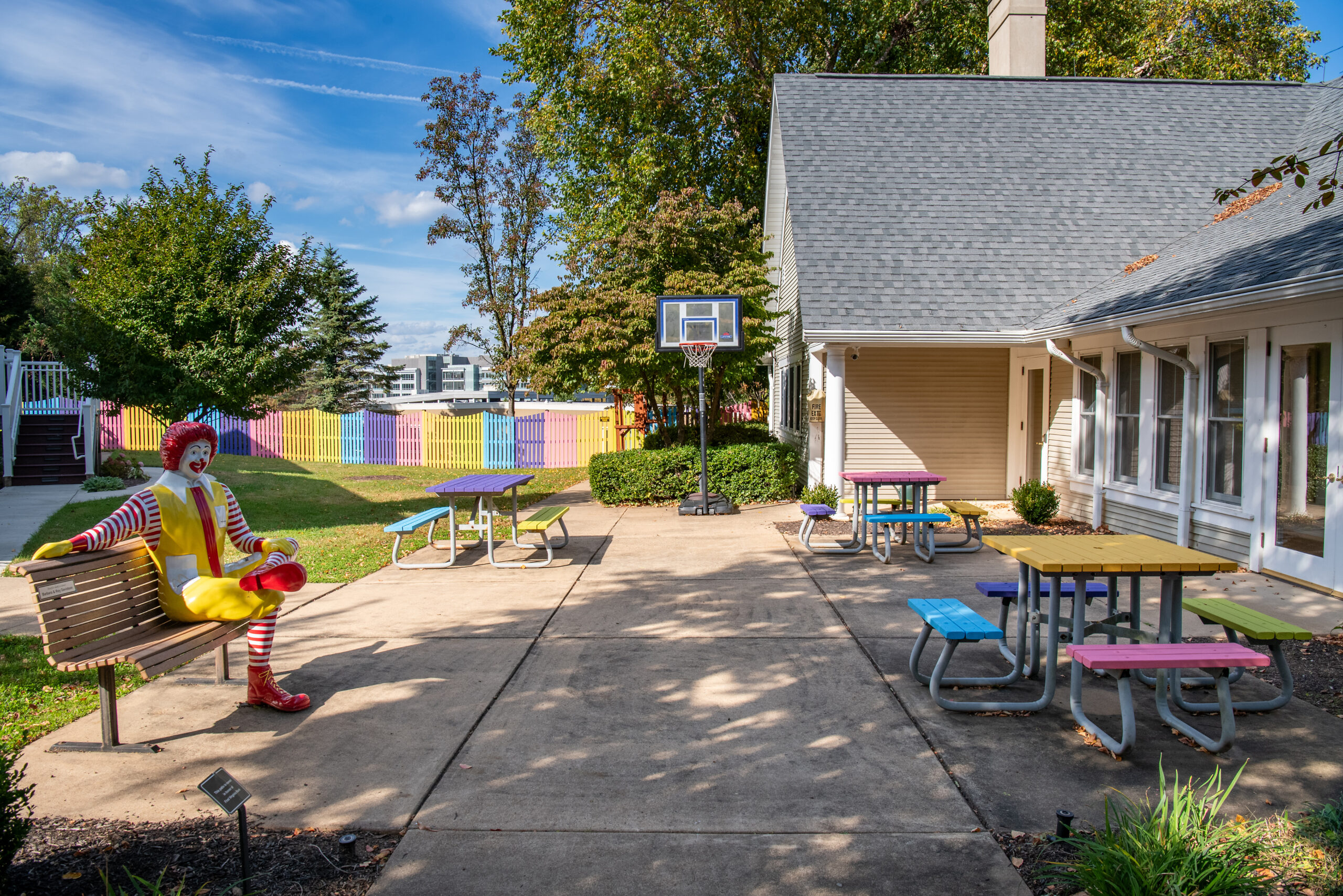 Outdoor seating area with colorful picnic tables and a Ronald McDonald statue on a bench. A basketball hoop is mounted on a wall. Trees and a multicolored fence are visible in the background.