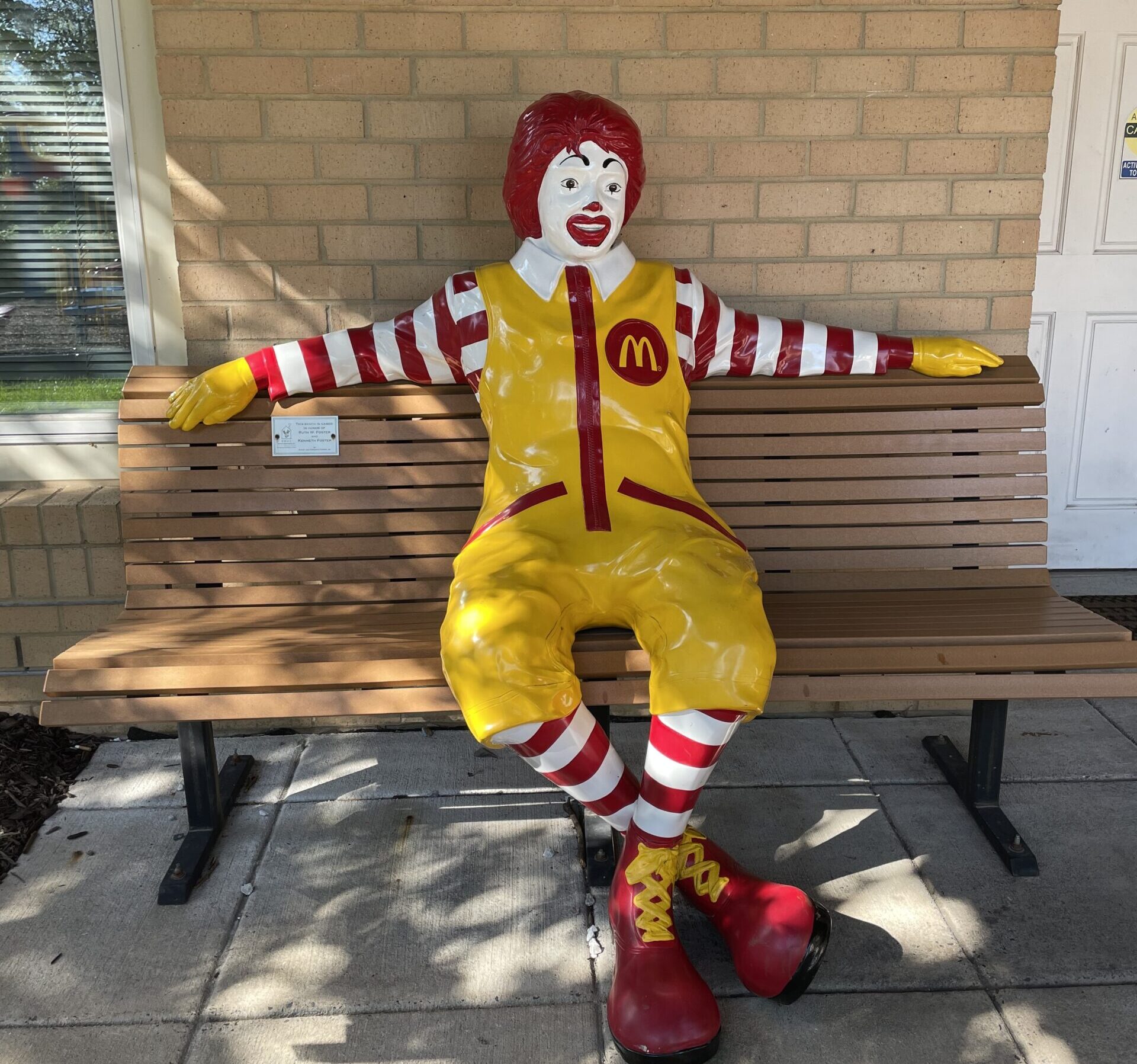 A Ronald McDonald statue is seated on a wooden bench outside a brick building. The statue is dressed in a yellow jumpsuit with red and white striped sleeves, red shoes, and a red wig.