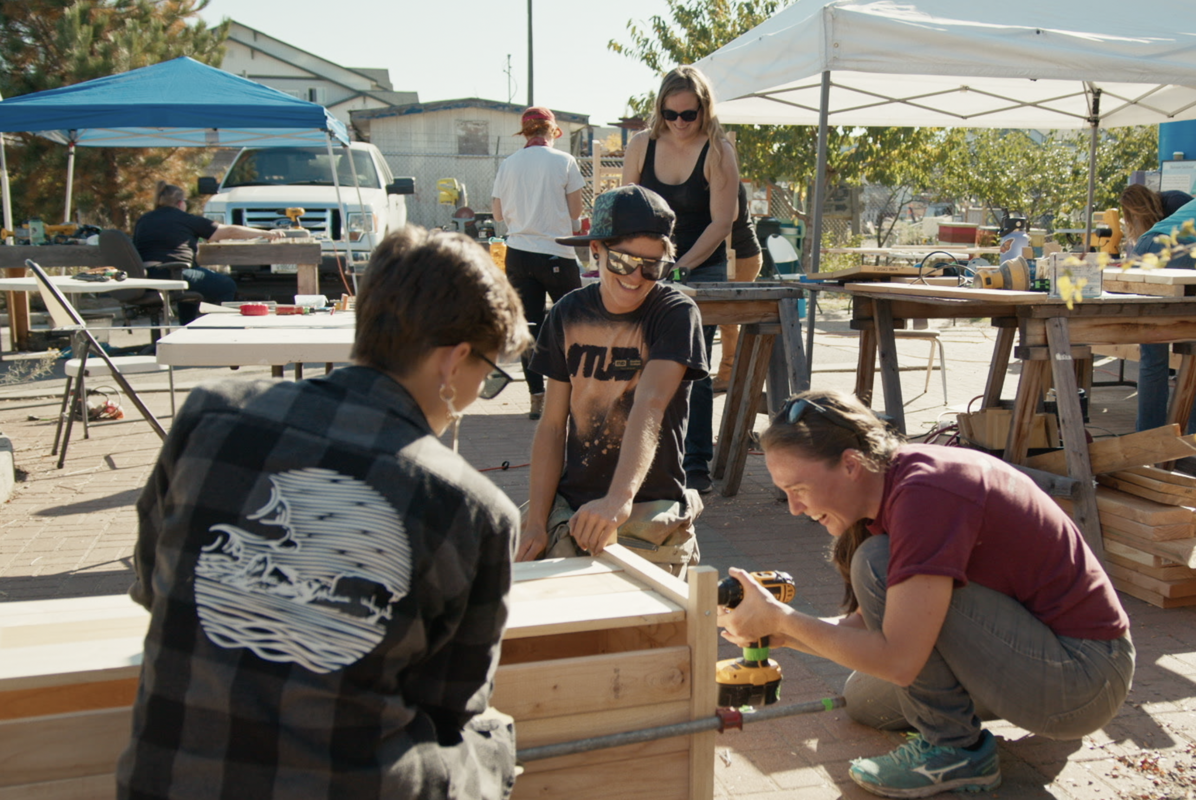A group of people work together on woodworking projects outdoors under tents, with various tools and wooden structures around them.