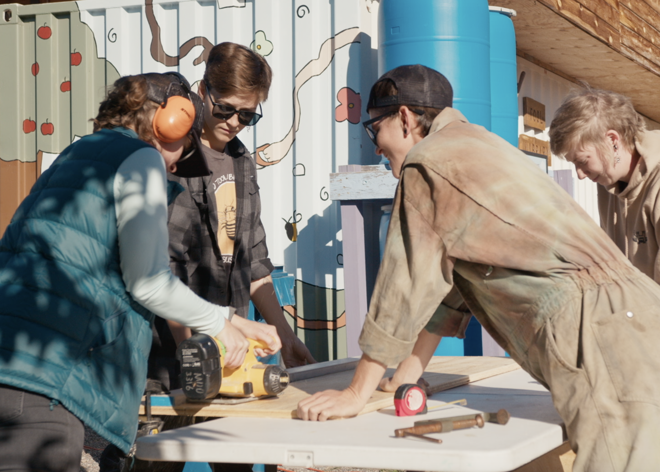 Three people are working on cutting a wooden plank with a power saw on a table outdoors. Another person is working in the background. Various tools and materials are on the table.